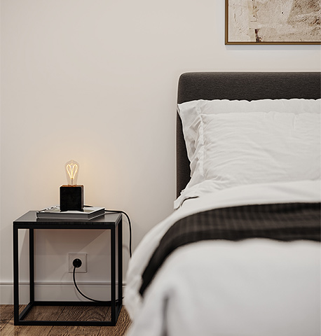 Nahaufnahme des Bettes und Beistelltisches in Schwarz mit einer Tischleuchte aus einer Glühbirne